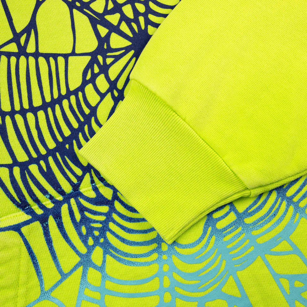 A close-up of the Sp5der Web Graphic Hoodie in neon green (440GSM heavyweight streetwear) shows the fabric texture and a bold, abstract spider web graphic in navy and light blue on the sleeve.