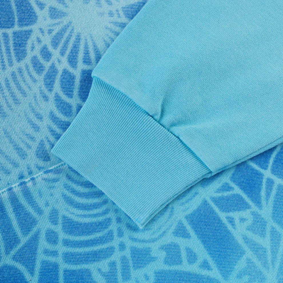A close-up of the Sp5der “Blue Venom” Web Hoodie – Sky Blue Fade, a 440GSM heavyweight oversized streetwear pullover, shows its sleeve resting on blue fabric with a white spider web pattern.