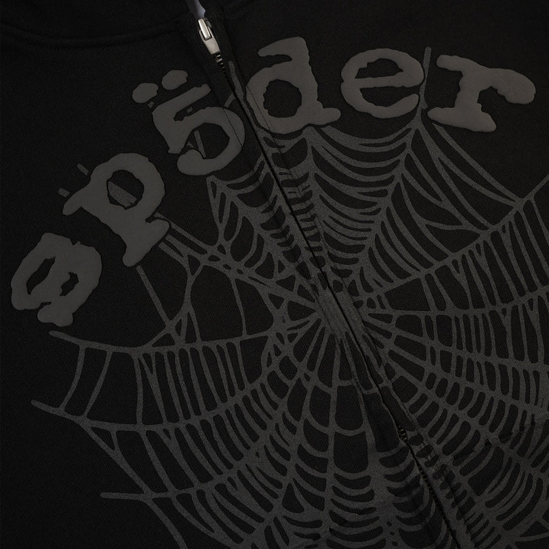 Close-up of the Sp5der Tonal Web Zip Hoodie – Black, a 440GSM heavyweight streetwear piece with raised, stylized "spider" lettering, a tonal web graphic across the fabric, and a partially visible zipper at the top center.