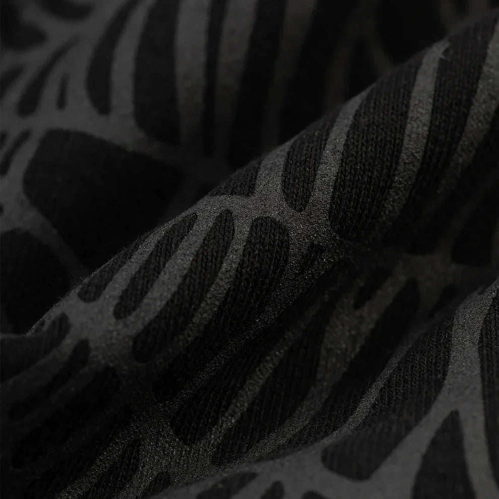 Close-up of the Sp5der Tonal Web Zip Hoodie in black 440GSM heavyweight fabric, showcasing a raised abstract pattern with organic shapes and a tonal spider web graphic. Lighting reveals matte and slightly shiny contrasts on the streetwear hoodie.
