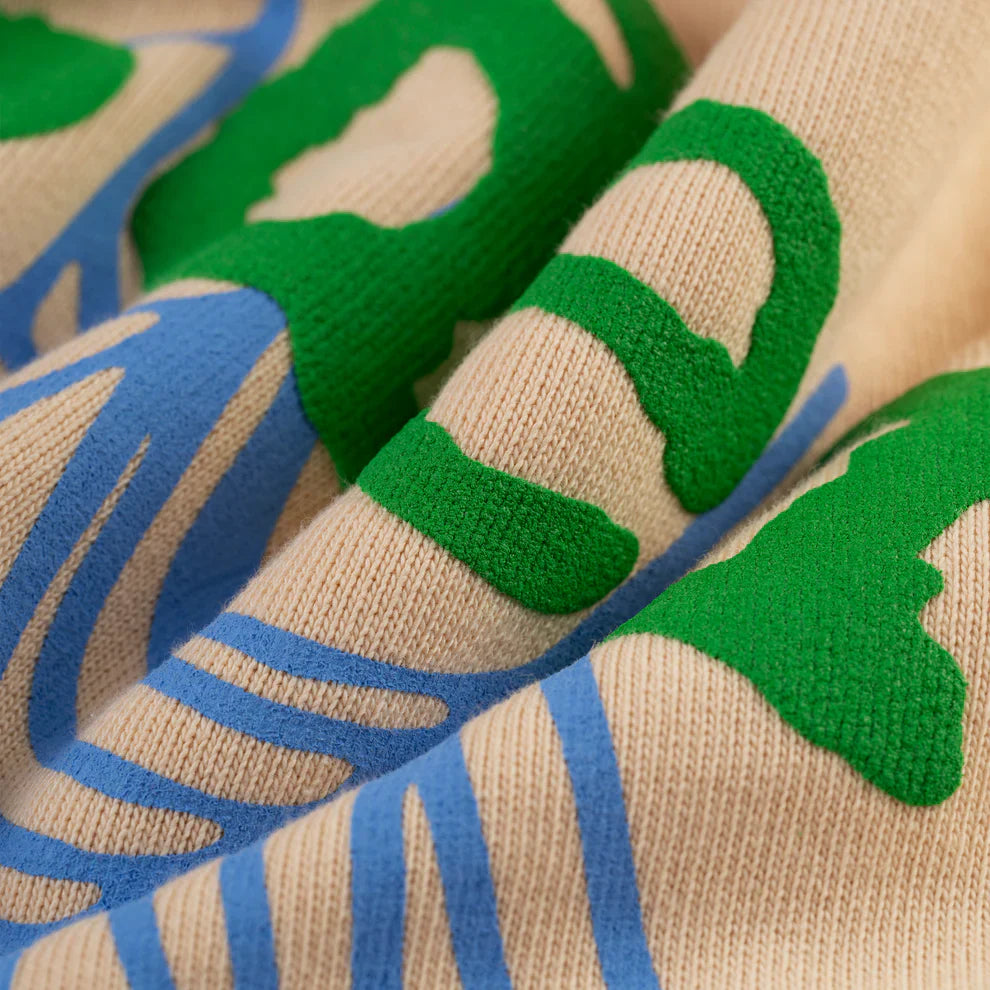 A close-up of heavyweight 440GSM cotton fleece from the Sp5der “Sky Web” Hoodie—Cream/Blue/Green—shows textured abstract designs and gentle folds, capturing its oversized Y2K streetwear style.