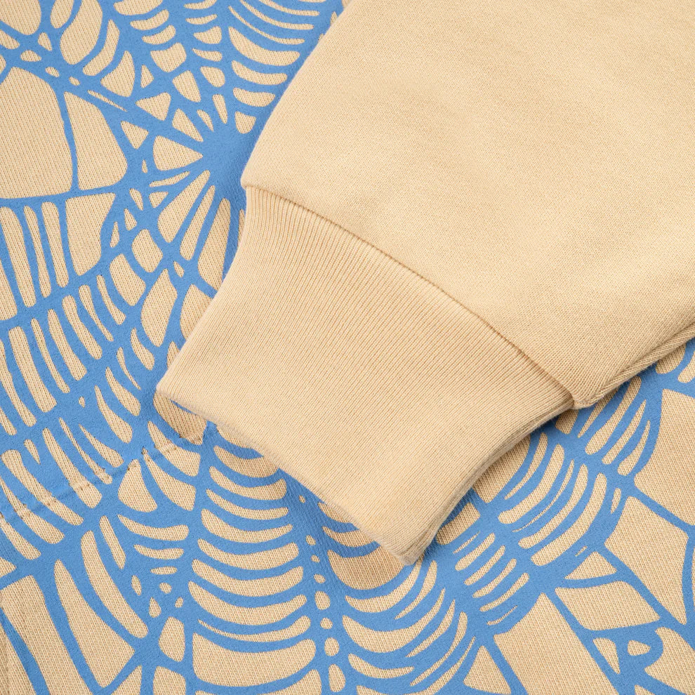 A sleeve of the Sp5der “Sky Web” Hoodie – Cream / Blue / Green 440GSM Oversized Streetwear Pullover rests on blue patterned fabric, its ribbed cuff and abstract web design highlighting Y2K streetwear style.