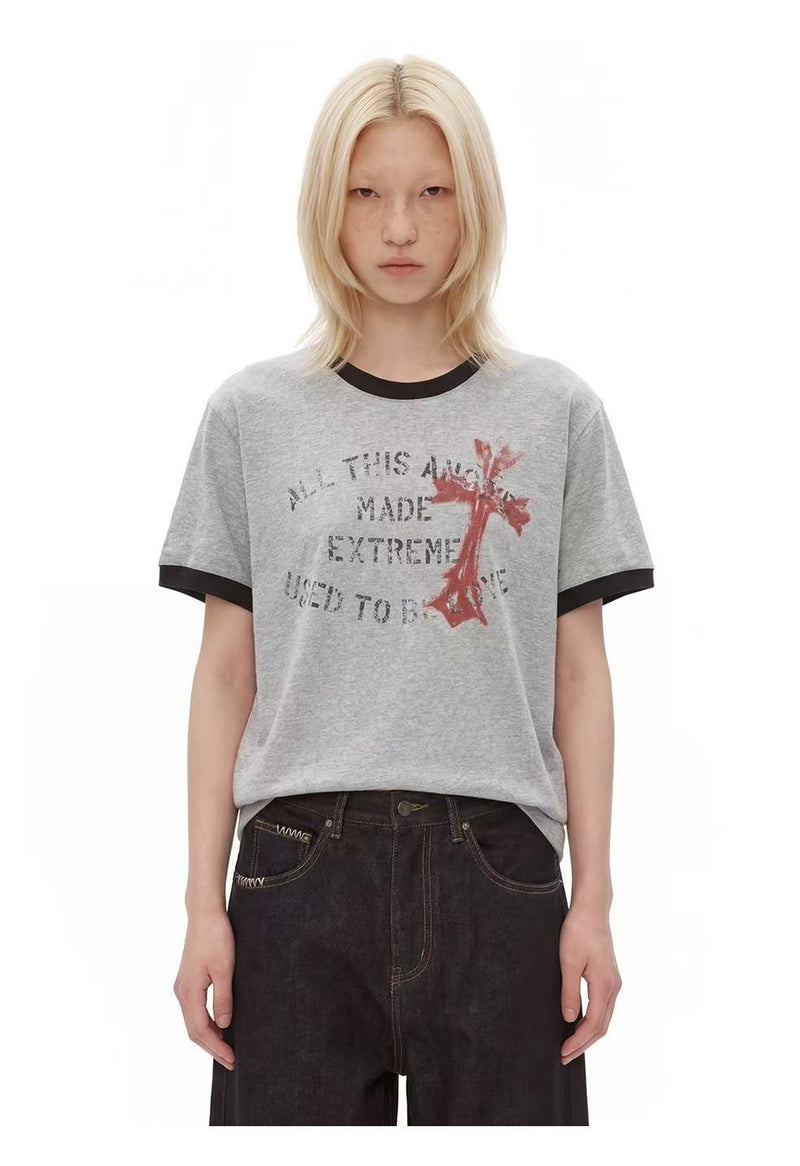 Model wearing a gray ringer T-shirt with distressed cross graphic and black trim, styled with dark jeans on a white background