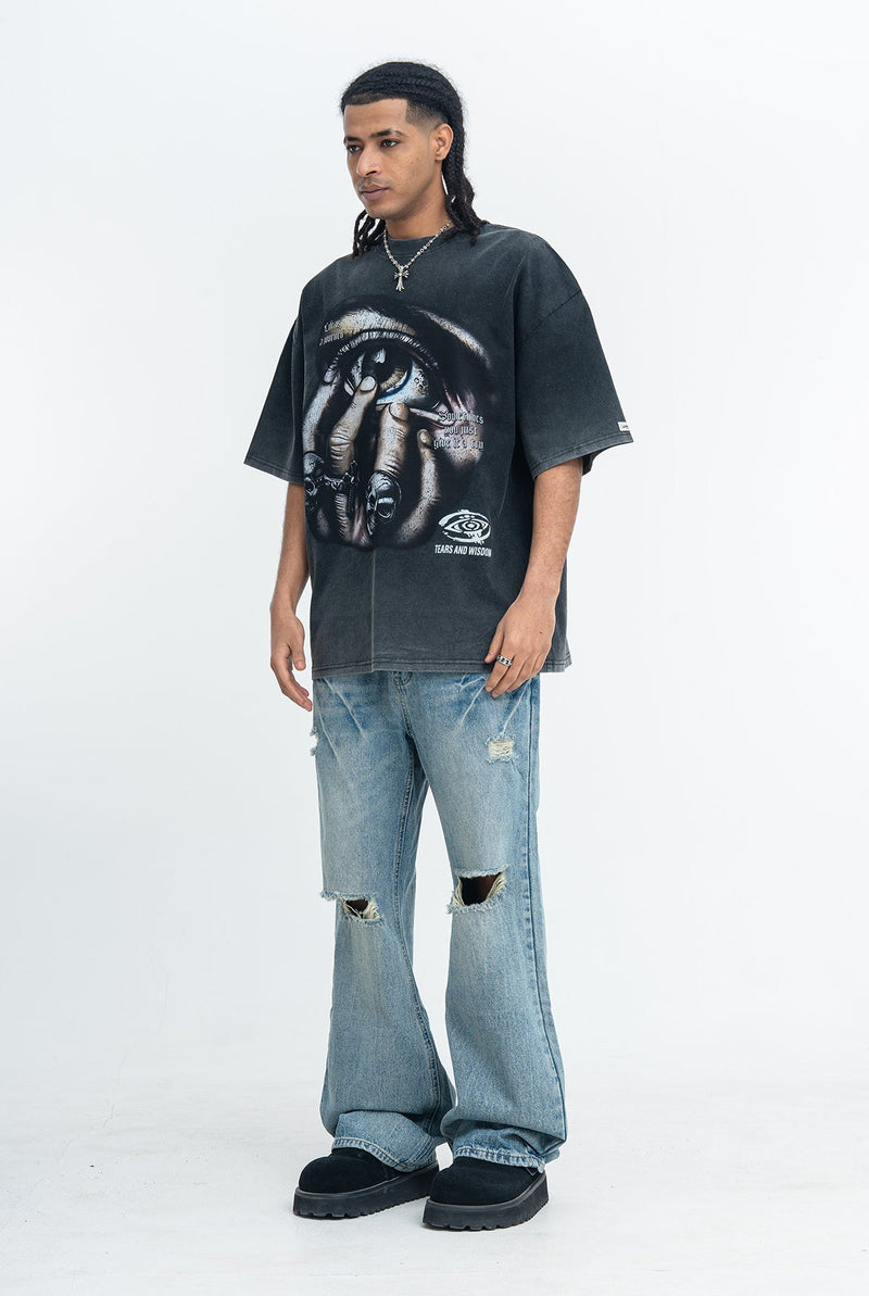 Oversized black graphic tee with eye print, paired with distressed baggy jeans and black shoes on a male model
