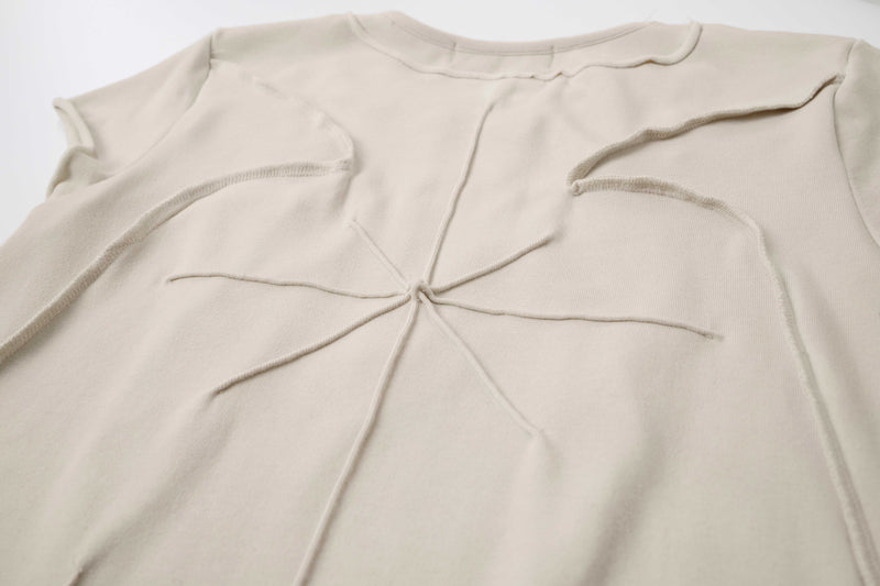 Beige sculpted cotton tee with raw seam paneling and abstract stitched detail on a minimalist white background