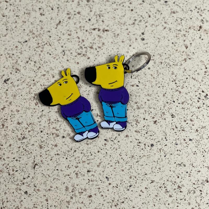 Two identical acrylic keychains from the &quot;Chill Guy Keychain Set – 10-Piece Meme-Inspired Collection&quot; feature a yellow, dog-like character with a purple shirt, blue pants, and white shoes on a speckled beige surface.