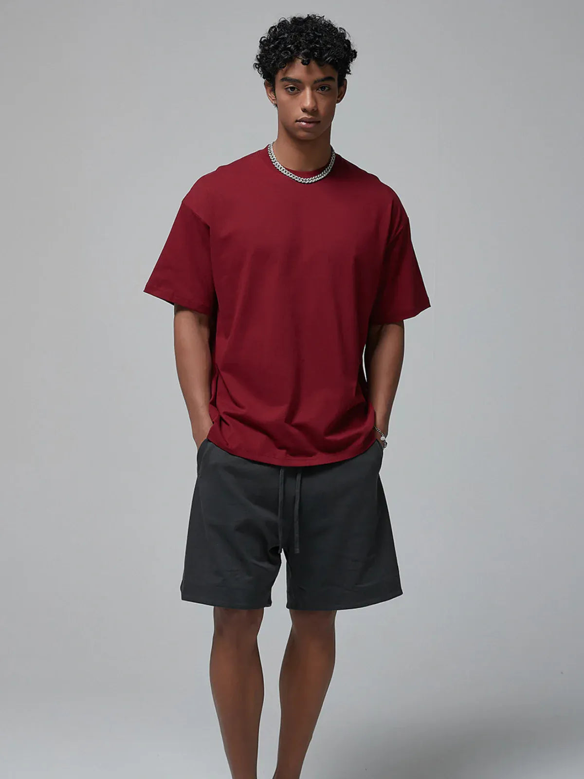 A man stands against a plain background wearing the Custom Anime Character Burgundy T-Shirt—Personalized 230GSM 100% cotton tee—with dark shorts and a necklace, his hands in his pockets.