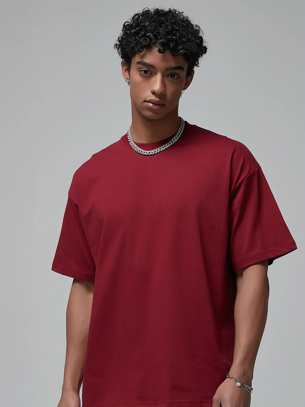 A person with curly hair, wearing a thick chain necklace and a Custom Anime Character Burgundy T-Shirt - Personalized 230GSM 100% Cotton Tee, stands against a plain gray background.