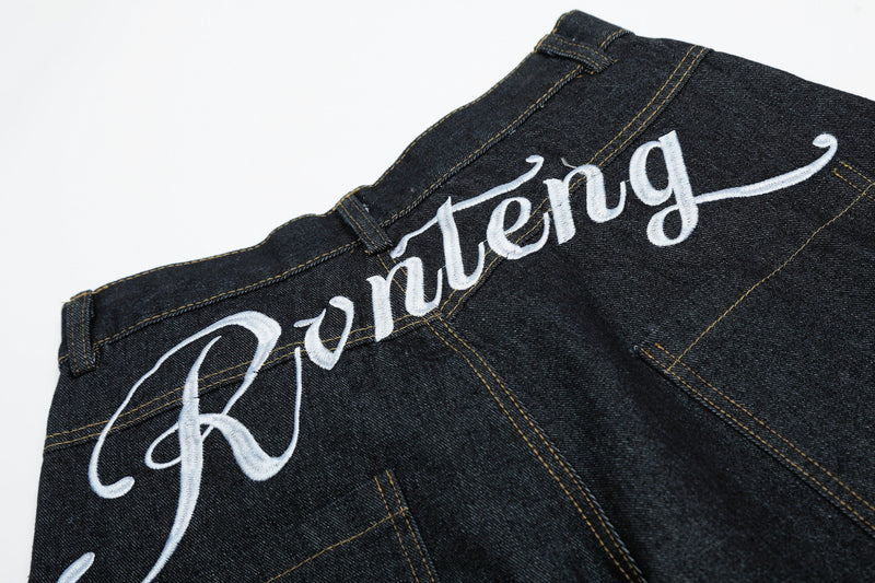 Back waist close-up of black denim jeans showing white script embroidery across the yoke