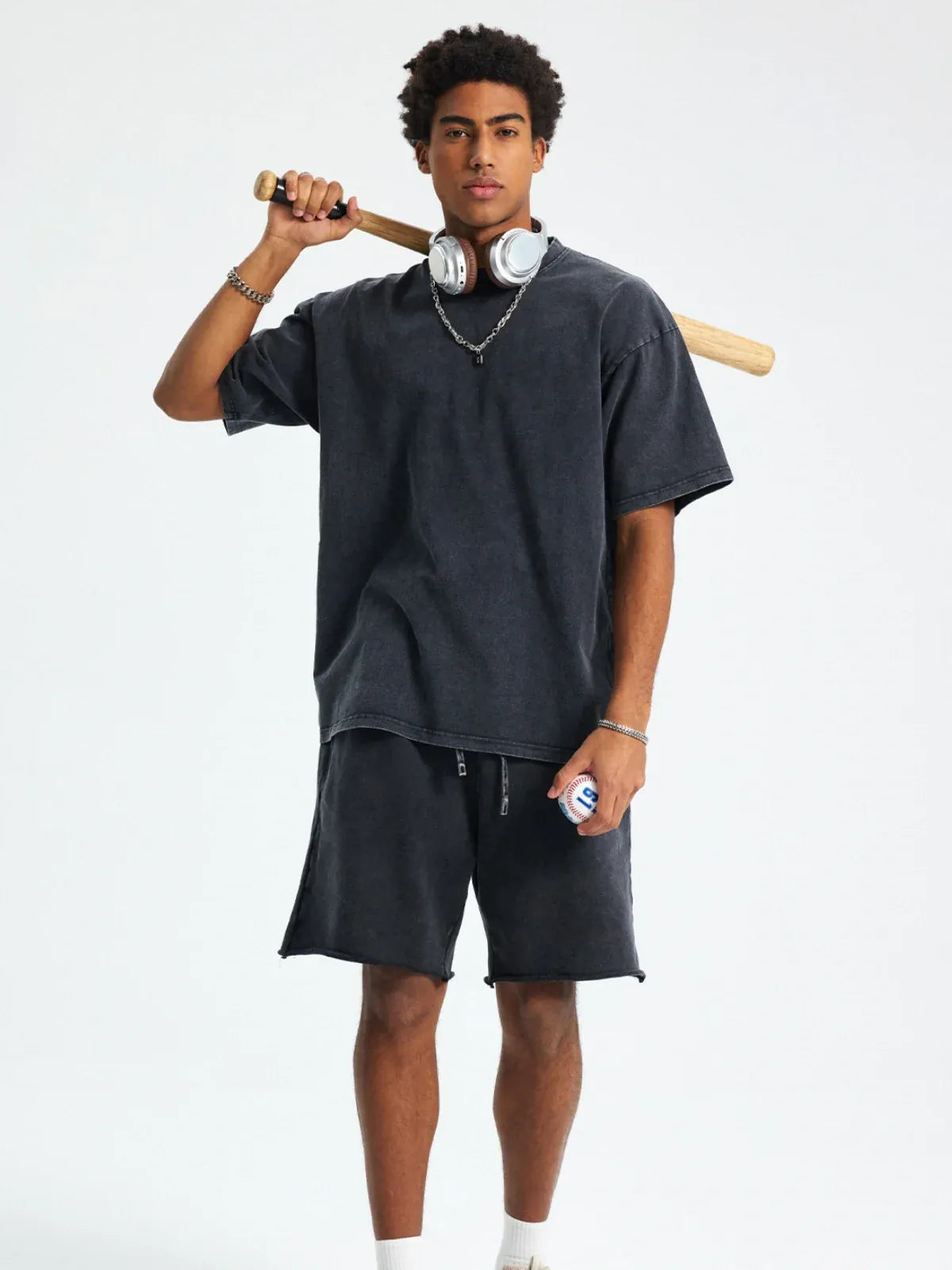 A young man stands against a plain background, holding a baseball bat and ball, wearing the Custom Anime Character Black Washed T-Shirt – Personalized Anime Style Tee, with headphones around his neck and shorts.