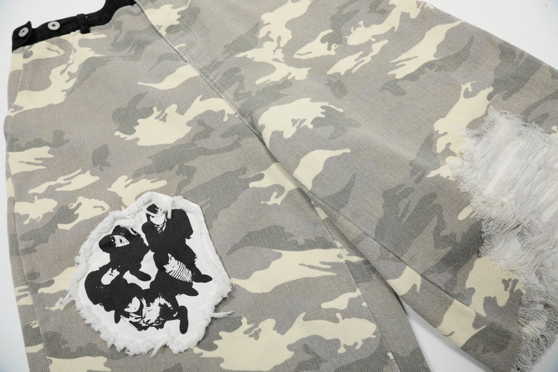 Angle view of gray camouflage jorts showing front graphic patch and a large ripped leg opening