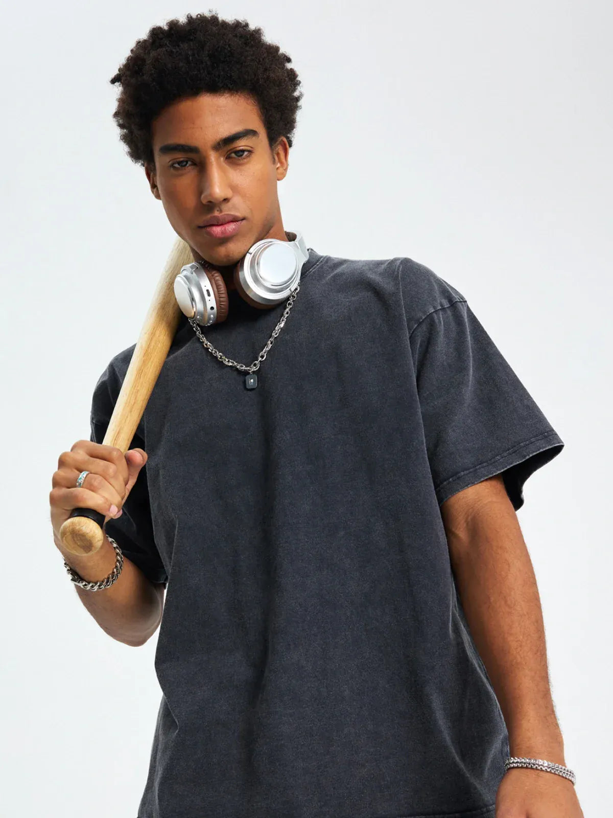 Young man wearing a Custom Anime Character Black Washed T-Shirt with headphones around his neck holds a baseball bat over his shoulder, looking at the camera against a plain background.