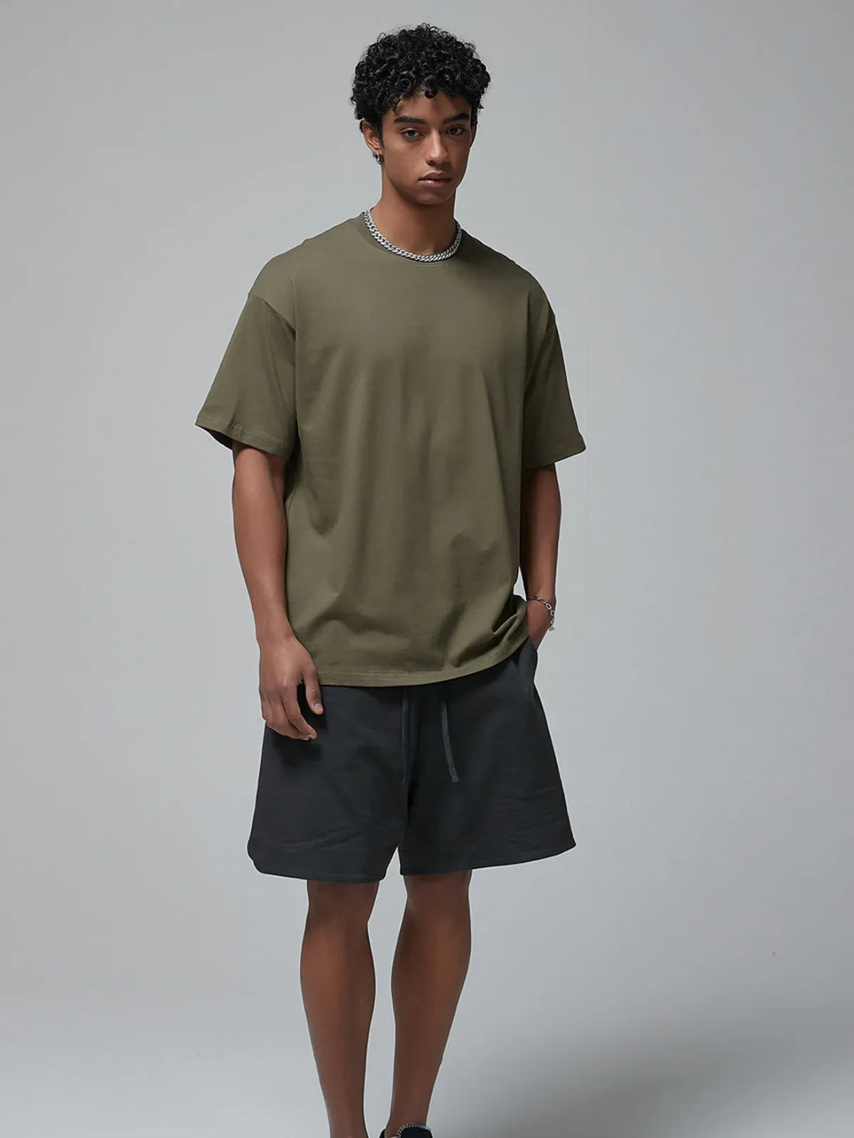 A man stands against a plain background wearing a Custom Anime Character Olive Green T-Shirt - Personalized 230GSM 100% Cotton Tee, paired with black shorts, a chain necklace, and a bracelet.