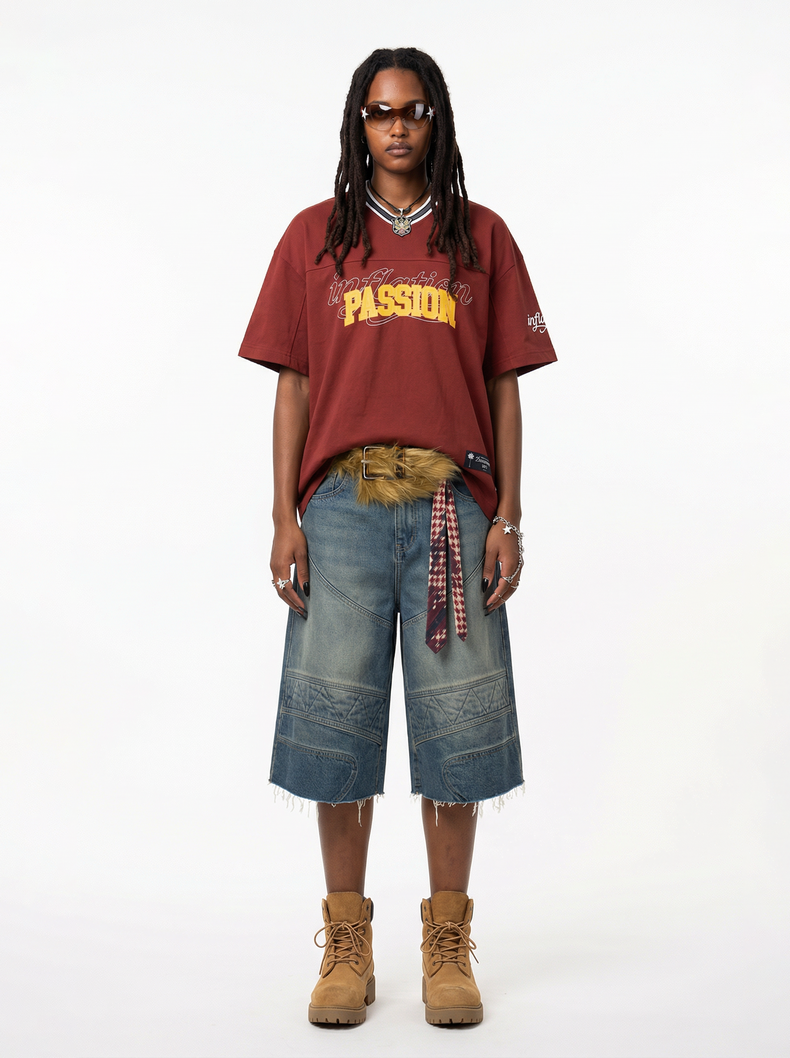 Model wearing a burgundy graphic V-neck tee with oversized denim shorts, sunglasses, and tan lace-up boots