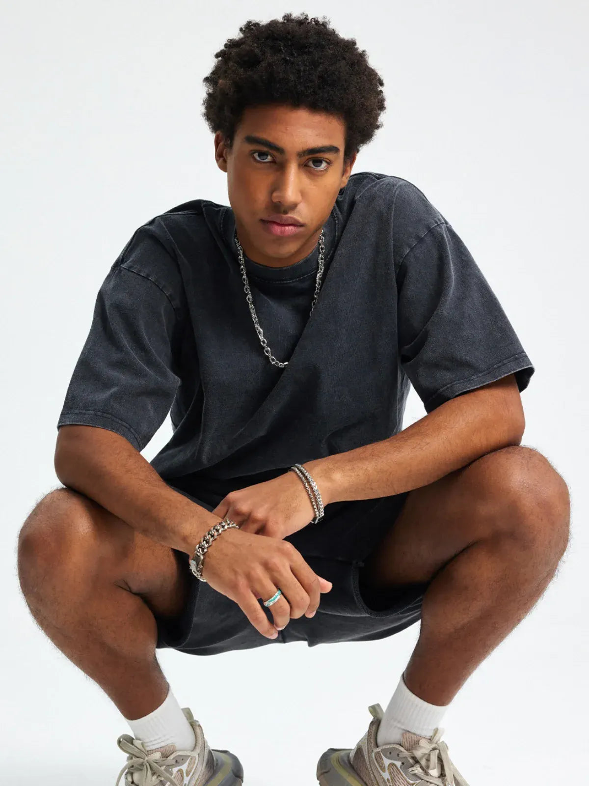 A young man squats against a plain background, wearing the Custom Anime Character Black Washed T-Shirt - Personalized Anime Style Tee, paired with shorts, sneakers, and jewelry.