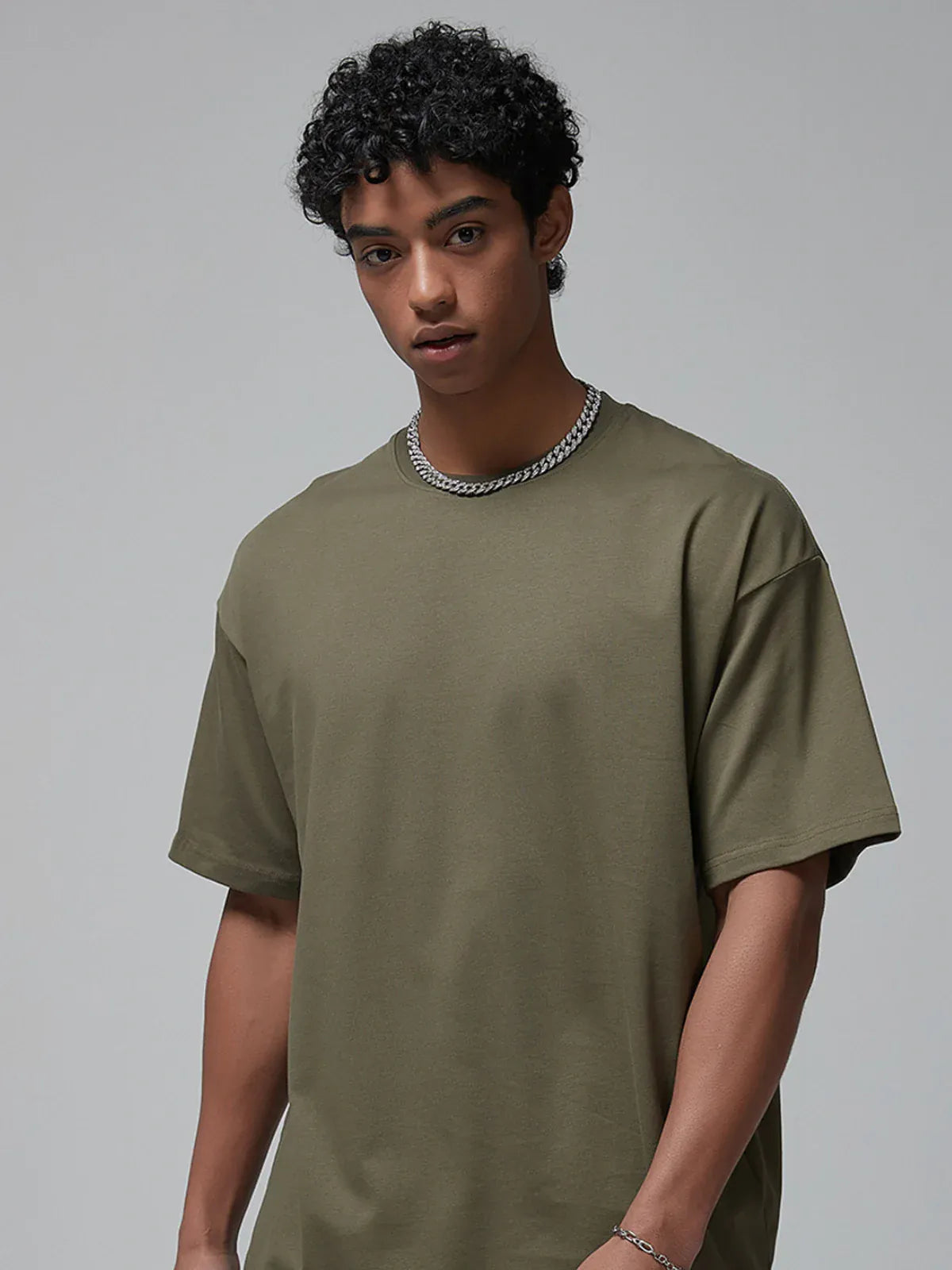 A young man with curly hair in a Custom Anime Character Olive Green 100% Cotton T-Shirt (230GSM) and a silver chain necklace stands against a plain gray background.