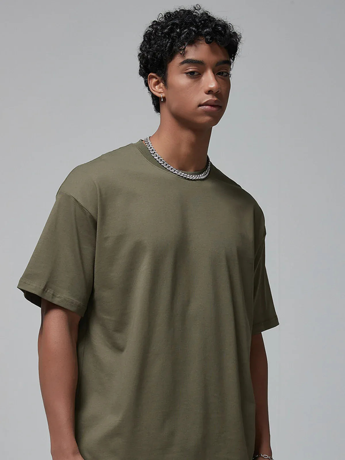 A person with short curly hair wears the Custom Anime Character Olive Green T-Shirt – a personalized 230GSM 100% cotton tee – and a silver chain necklace, standing against a plain light gray background.