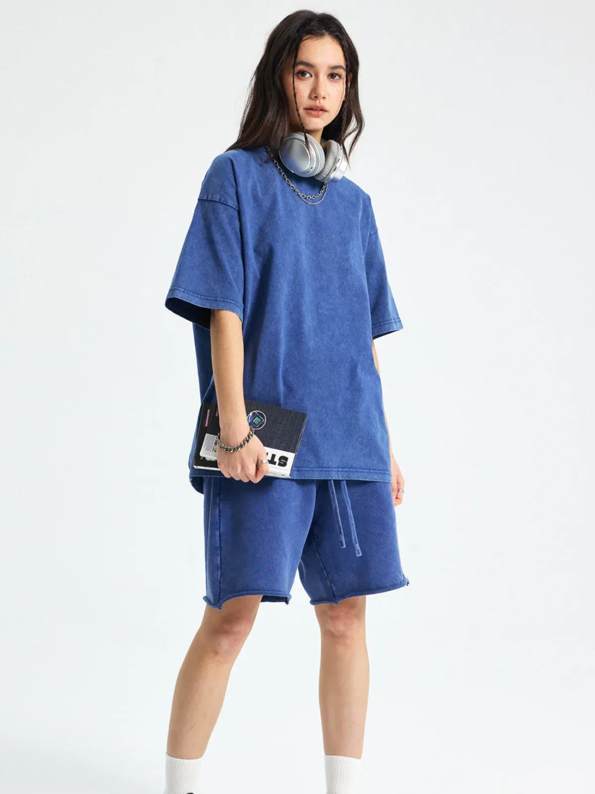 A person stands in a Custom Anime Character Periwinkle Blue Washed T-Shirt - Personalized Anime Style Tee and matching shorts, holding a notebook with headphones around their neck, against a plain light background.