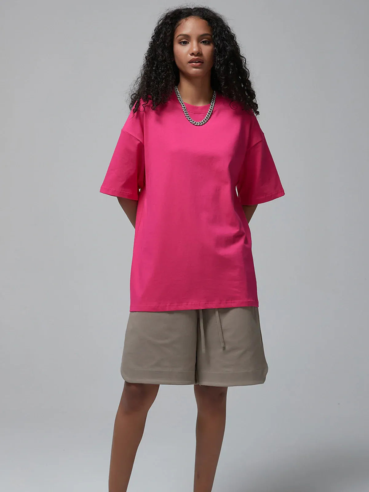 A person stands against a plain background, wearing the Custom Anime Character Hot Pink T-Shirt – a personalized 230GSM 100% cotton tee – along with beige knee-length shorts and a silver chain necklace.