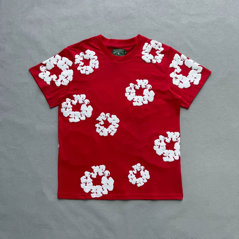 The Denim Tears Red Cotton Wreath Allover Print T-Shirt lies flat on a gray surface, showcasing white wreath-inspired graphics across the front and sleeves.