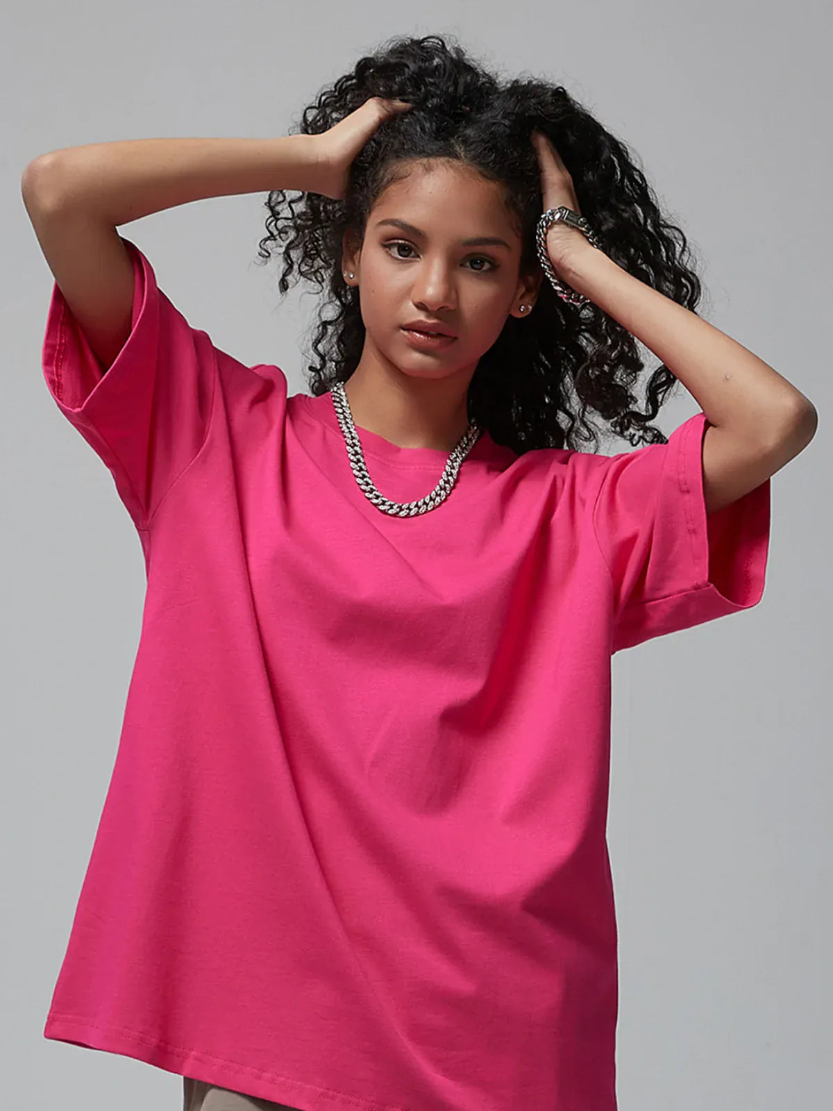 A young woman with curly hair wears a Custom Anime Character Hot Pink T-Shirt—Personalized 230GSM 100% Cotton Tee—and a chunky chain necklace, posing with both hands in her hair against a plain background.