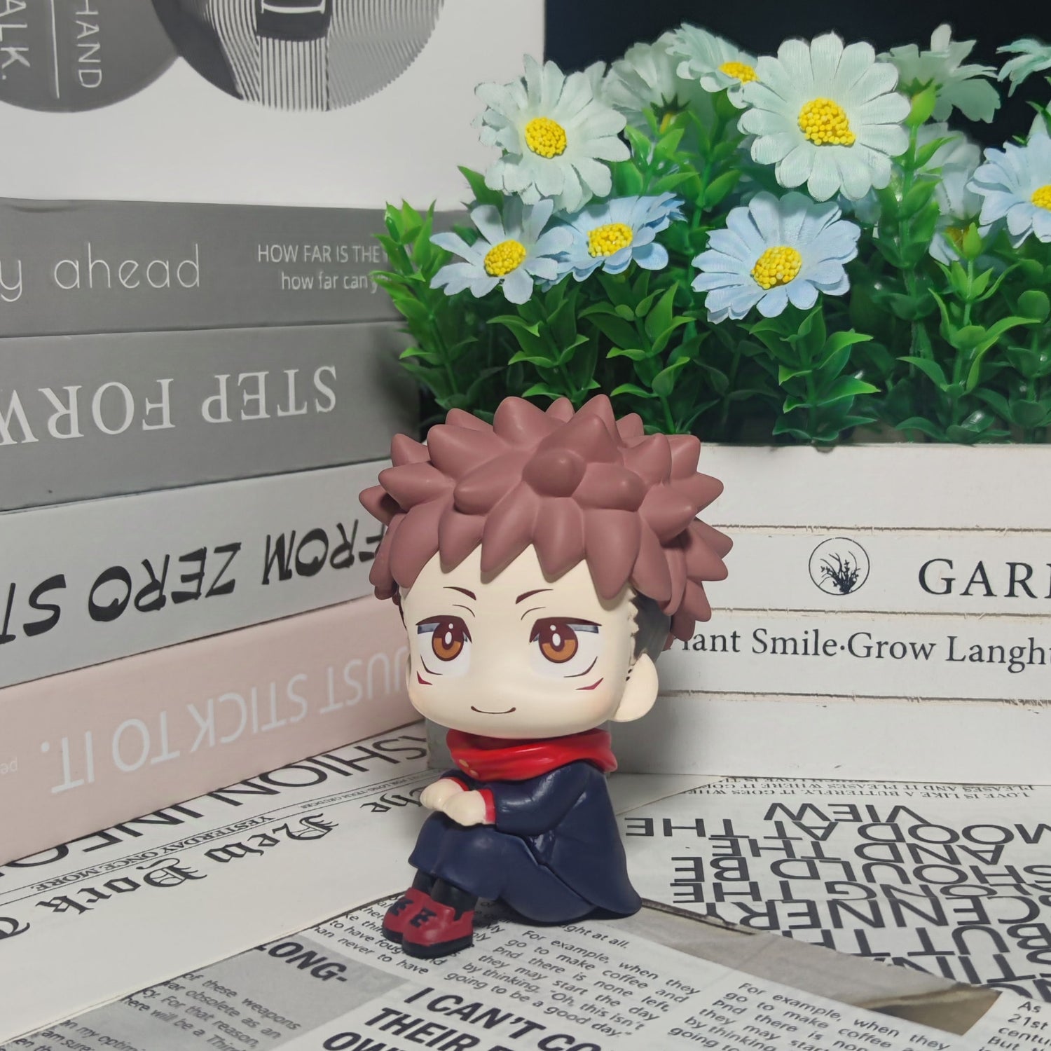 The Itadori Yuji Anime Figure, a 12cm Jujutsu Kaisen collectible with spiky brown hair and a red scarf, sits on newspaper print. With books and flowers in the background, it&