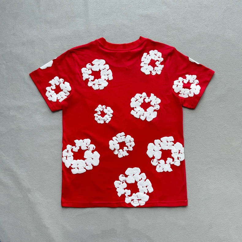 The Denim Tears Red Cotton Wreath Allover Print T-Shirt lies flat on a grey surface, with multiple white wreath graphics scattered across the back.