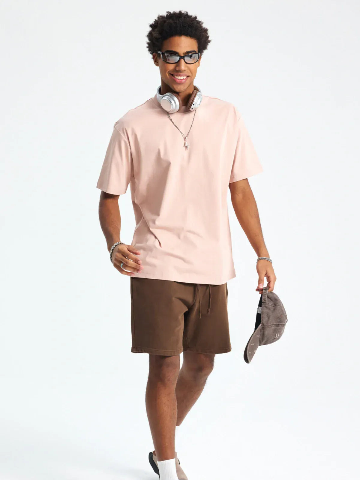A man wearing the Custom Anime Character Blush Pink T-Shirt – a personalized 230GSM 100% cotton tee – along with sunglasses and brown shorts, holds a gray cap while walking and smiling against a plain background.