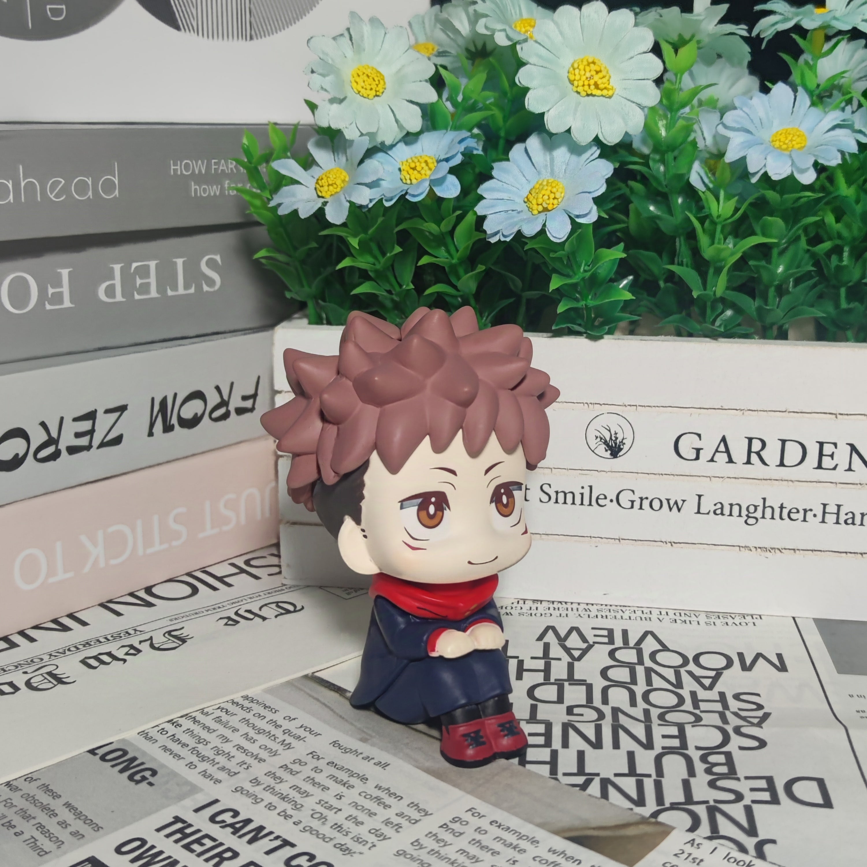 The 12cm Itadori Yuji Anime Figure from Jujutsu Kaisen, featuring a chibi-style pose, sits among newspapers. Nearby, books rest next to a wooden &quot;Garden&quot; box filled with white and blue artificial flowers—an ideal setting for anime figure collectors.