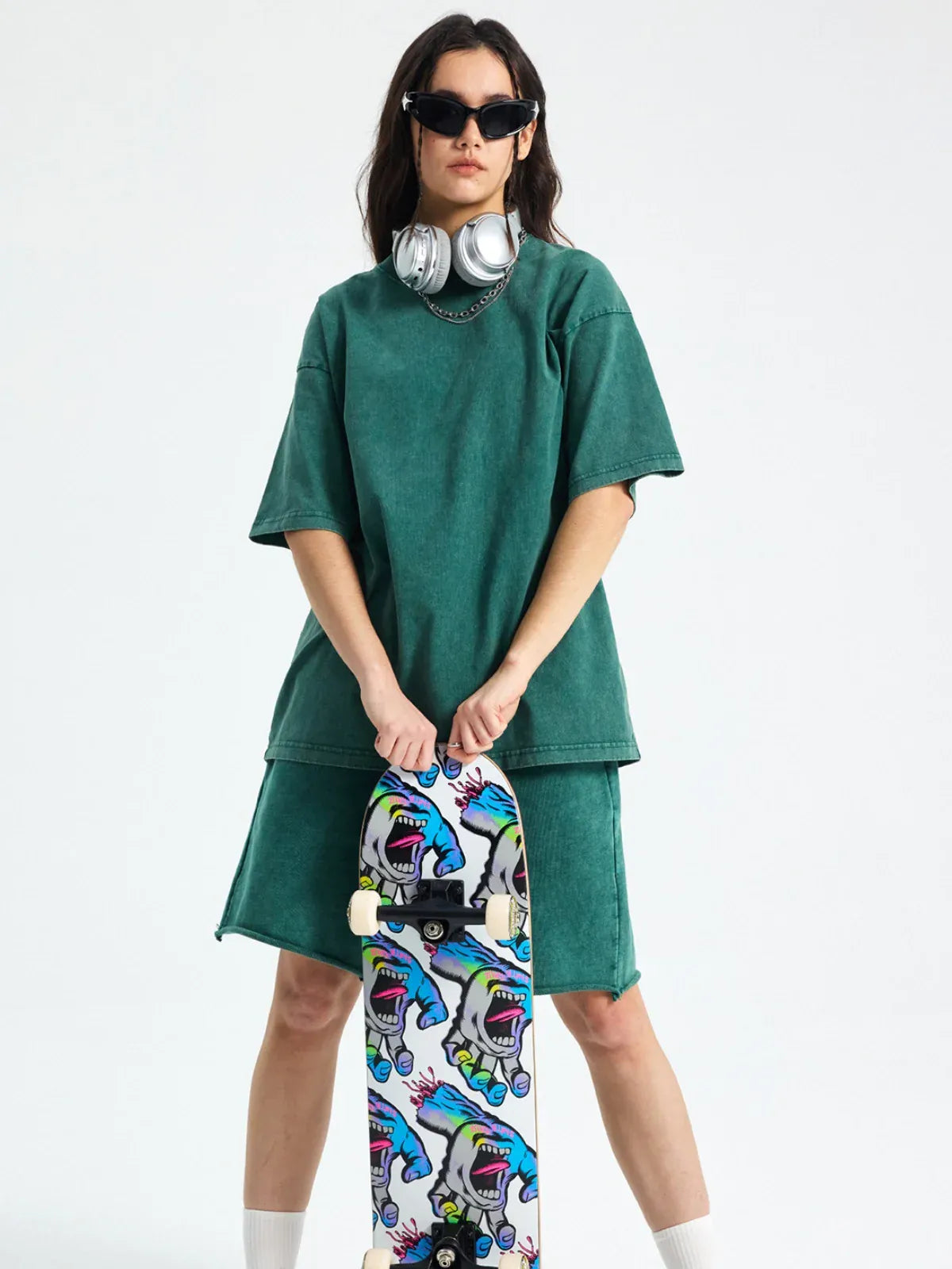 A person wearing the Custom Anime Character Green Washed T-Shirt stands holding a shark-themed skateboard, with headphones around their neck and sunglasses.