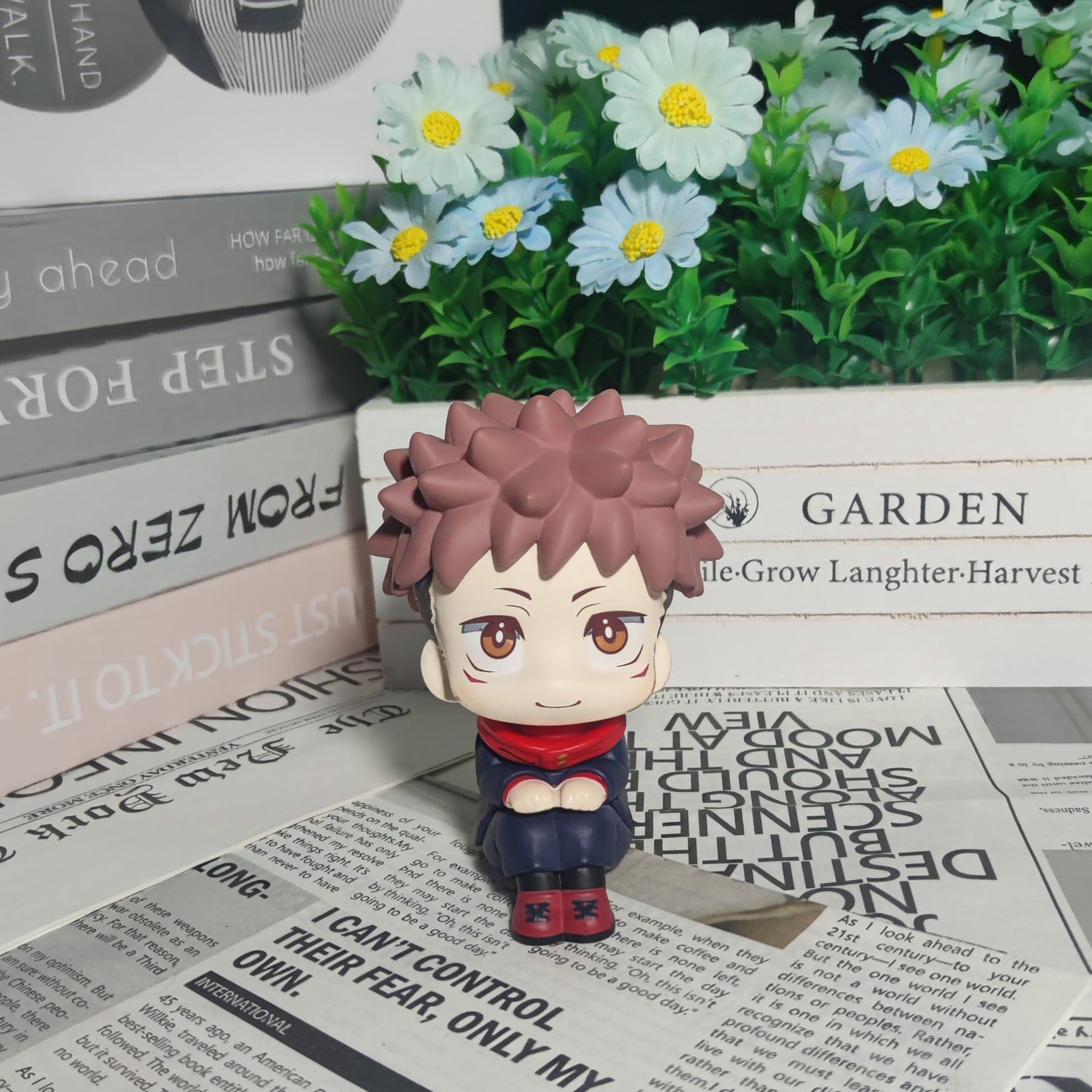 The Itadori Yuji Anime Figure by Jujutsu Kaisen features a cute, cross-legged pose with spiky brown hair and a red scarf. Displayed on newspapers with white and yellow flowers, as well as stacked books, it’s an enchanting collectible for anime figure enthusiasts.