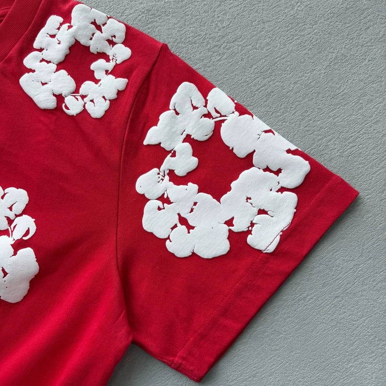 The Denim Tears Red Cotton Wreath Allover Print T-Shirt featuring large white abstract patterns is displayed flat on a gray textured surface.