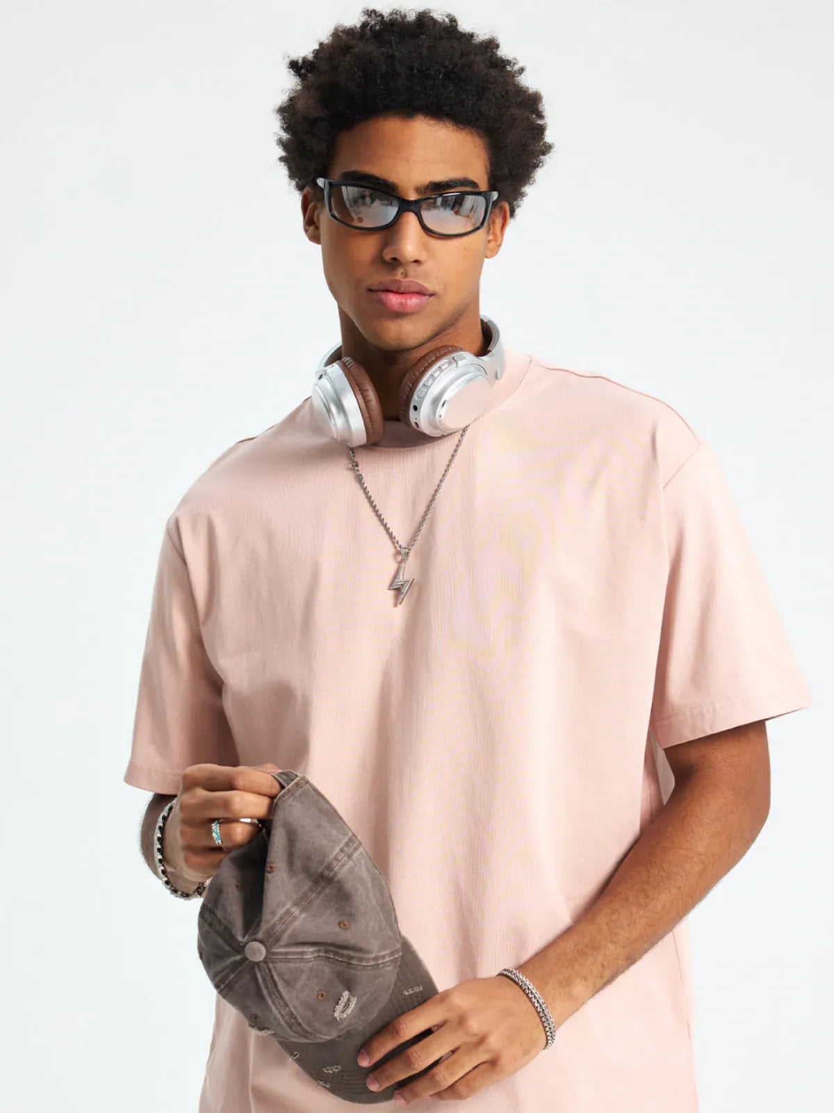 A young man in a Custom Anime Character Blush Pink T-Shirt (230GSM 100% Cotton), with sunglasses, headphones around his neck, and holding a gray baseball cap against a plain background.