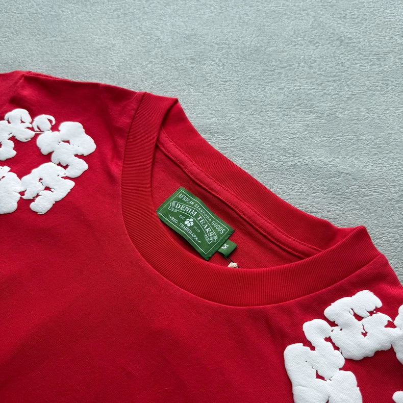 The Denim Tears Red Cotton Wreath Allover Print T-Shirt features white textured lettering near the shoulders, a green DENIM TEARS collar label, and is displayed laid flat on a light gray background.