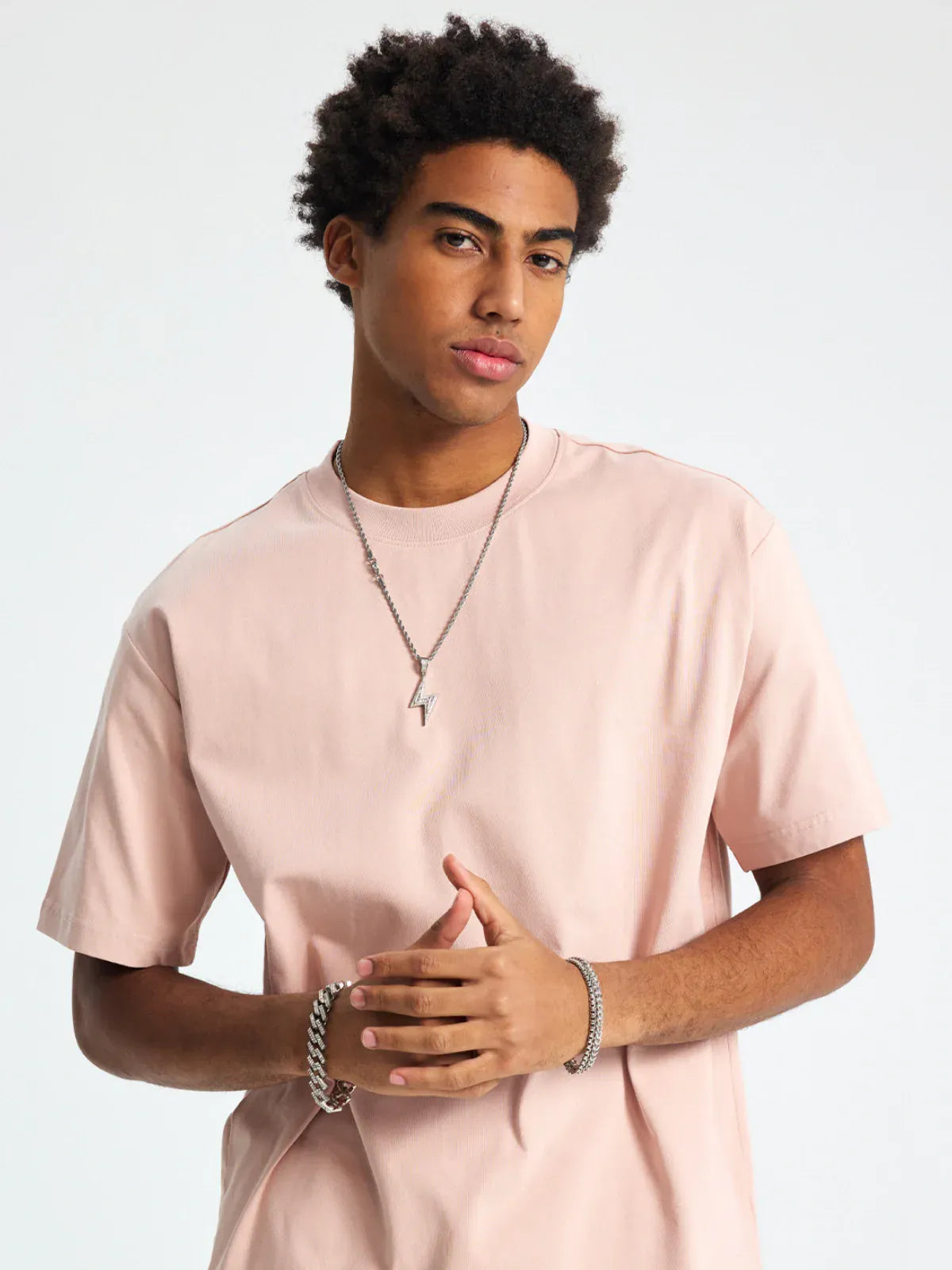 A person with short curly hair wears the Custom Anime Character Blush Pink T-Shirt – a personalized 230GSM 100% cotton tee – along with silver necklaces and a bracelet, standing against a plain light background.