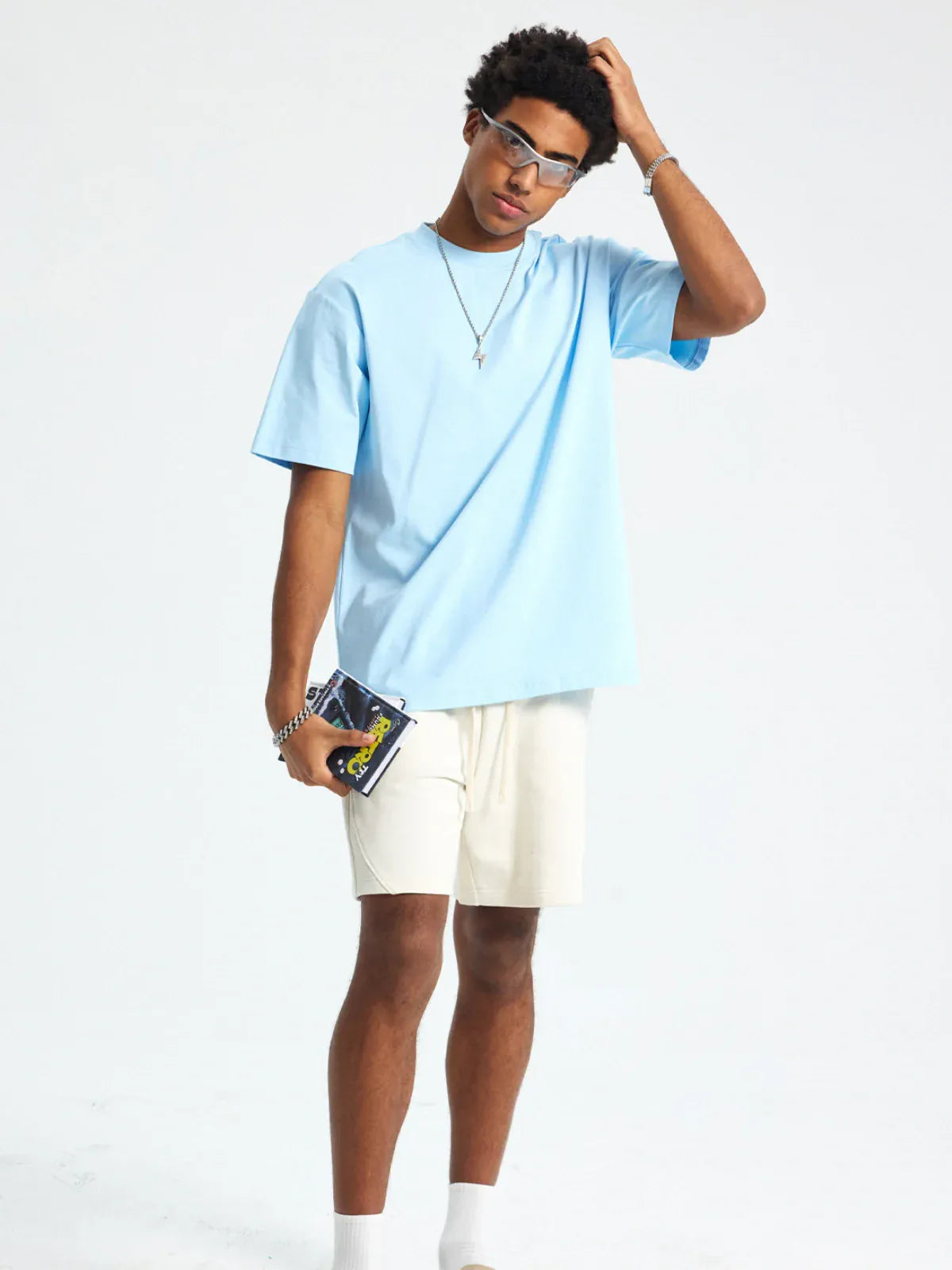 A person in a Custom Anime Character Sky Blue T-Shirt—Personalized 230GSM 100% Cotton Tee, beige shorts, and glasses stands against a plain background, holding a DVD and touching their head.