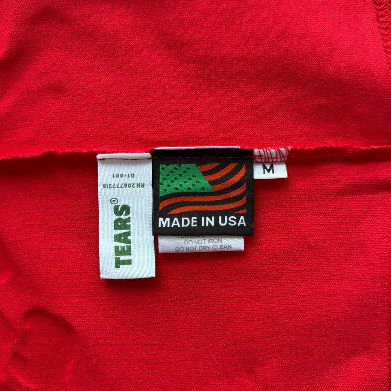 White tags on the Denim Tears Red Cotton Wreath Allover Print T-Shirt show TEARS®, a green and orange flag, MADE IN USA, care info, and an M tag for size medium.