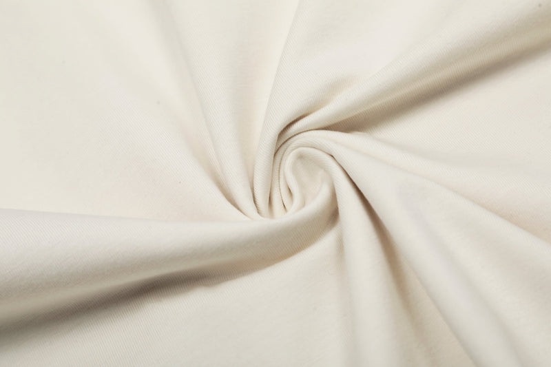 Cream cotton t-shirt fabric with soft spiral folds, shown in a close-up textured coastal tee backdrop