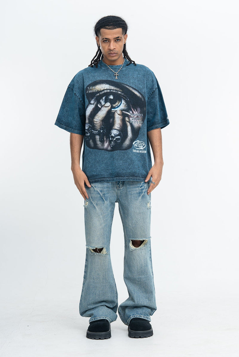 Model wearing an oversized blue graphic tee with a surreal eye print and baggy distressed jeans in a dark streetwear look