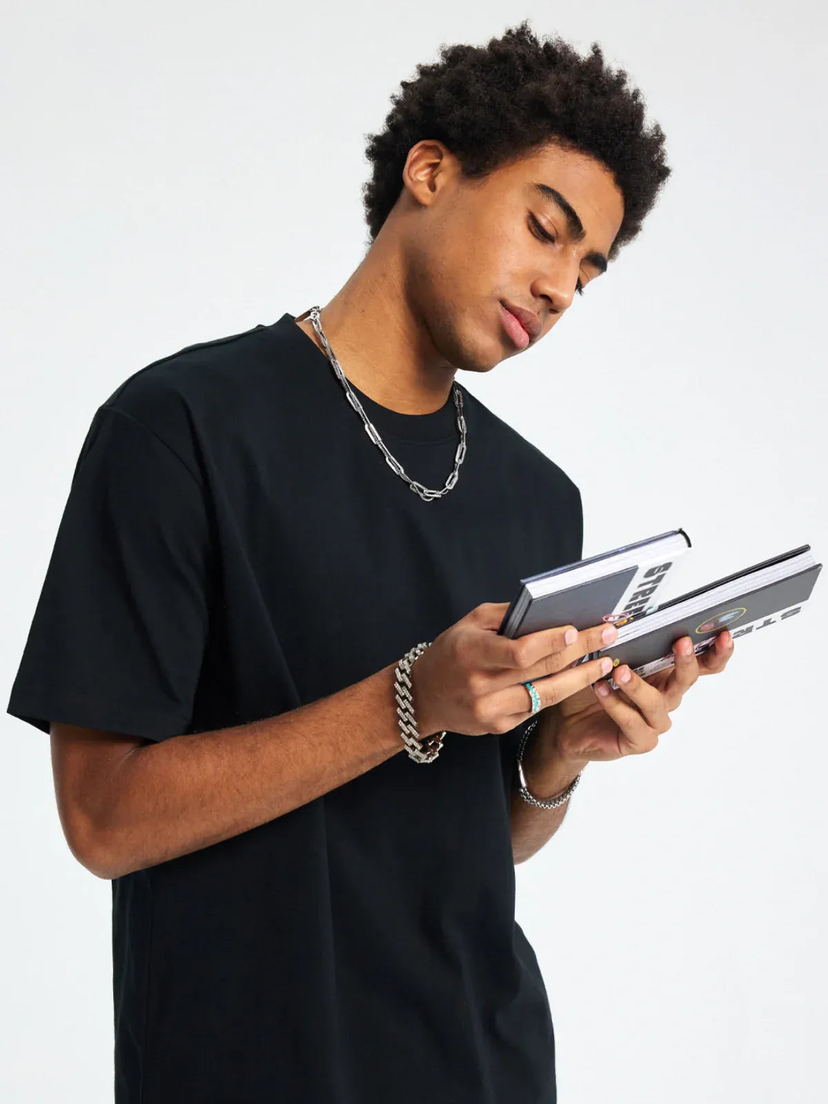 Wearing a Custom Anime Character Black T-Shirt - Personalized 230GSM 100% Cotton Tee and chain jewelry, a person looks down while flipping through a small book or notebook.