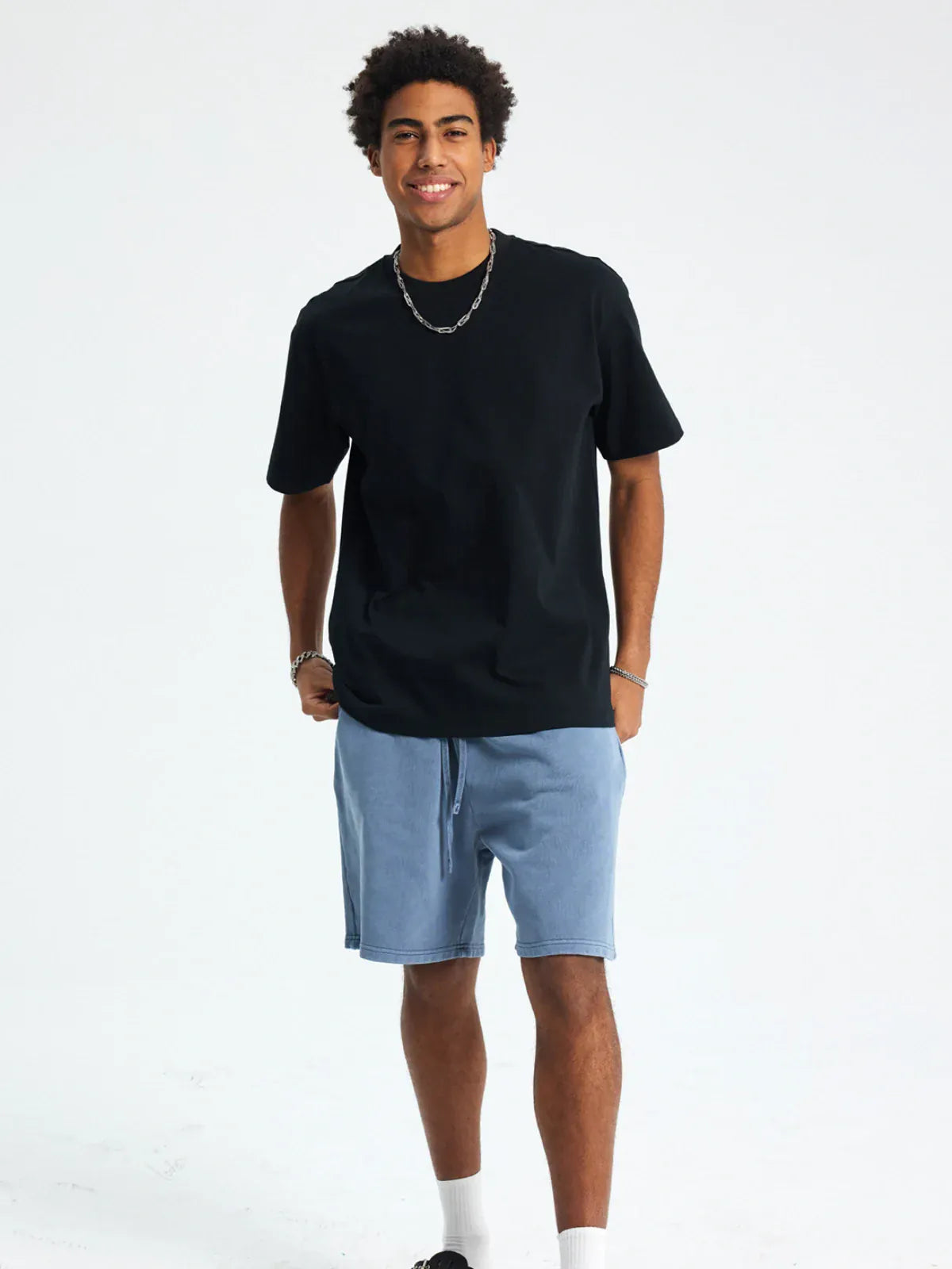 A man smiles with one hand in his pocket, wearing a Custom Anime Character Black T-Shirt - Personalized 230GSM 100% Cotton Tee, light blue shorts, a silver chain, and bracelets against a plain background.