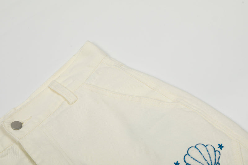 Close-up of cream cargo pants upper front with waistband, snap, and shell embroidery