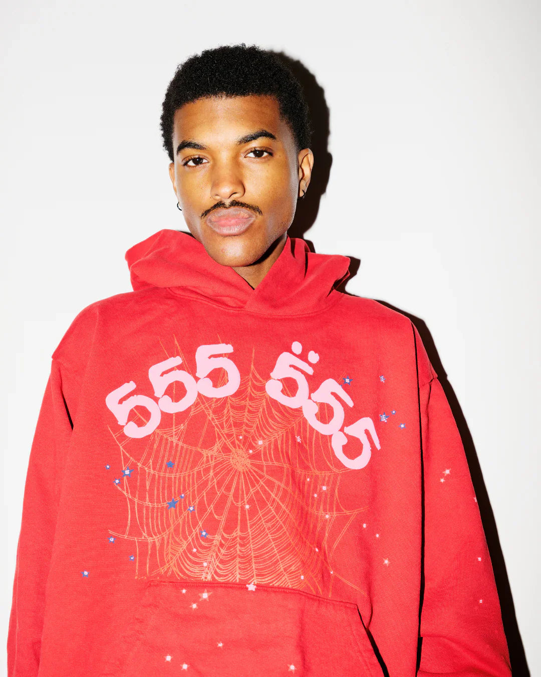 A young man stands against a plain white background, wearing the Sp5der “555” Web Hoodie – Red & Pink 440GSM Heavyweight Oversized Streetwear Pullover.