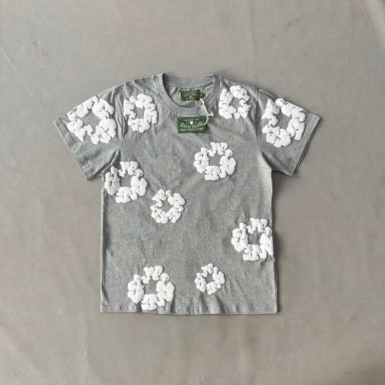 Denim Tears Grey Cotton Wreath Allover Print T-Shirt - 2 laid flat on a gray surface, features white allover wreath patterns and a green tag with white text attached near the collar.