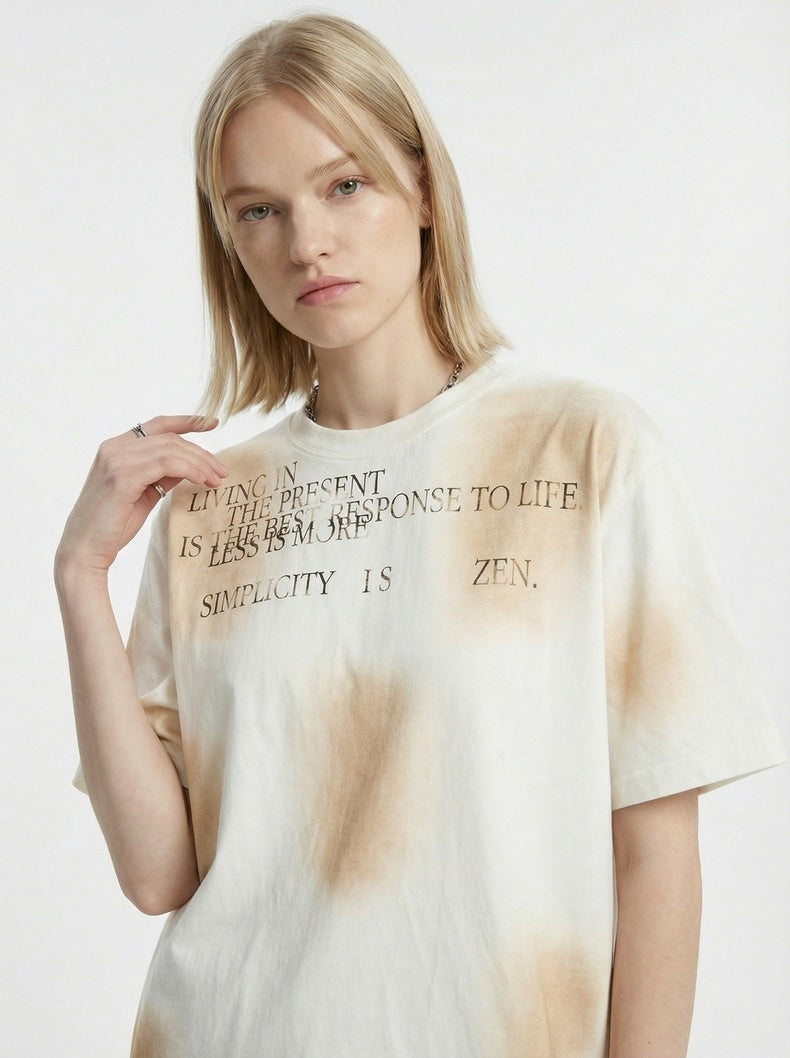 Cream spray-dyed vintage tee with minimalist text print, worn by a model in a relaxed streetwear style