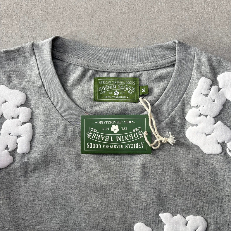 The Denim Tears Grey Cotton Wreath Allover Print T-Shirt - 2 features heather grey cotton, white puff-print designs at the shoulders, a white wreath graphic, a green collar label, and a matching green tag attached with string.