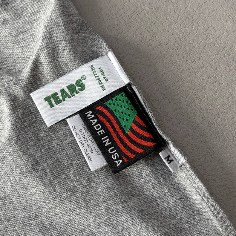 Close-up of a heather grey Denim Tears Grey Cotton Wreath Allover Print T-Shirt - 2 collar showing three tags: TEARS®, MADE IN USA with a stylized flag, and an M tag for medium size.