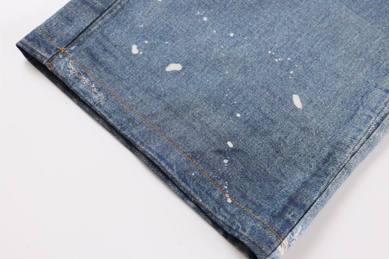 Close-up of lower pant leg on blue denim with white paint speckles and stitched hem