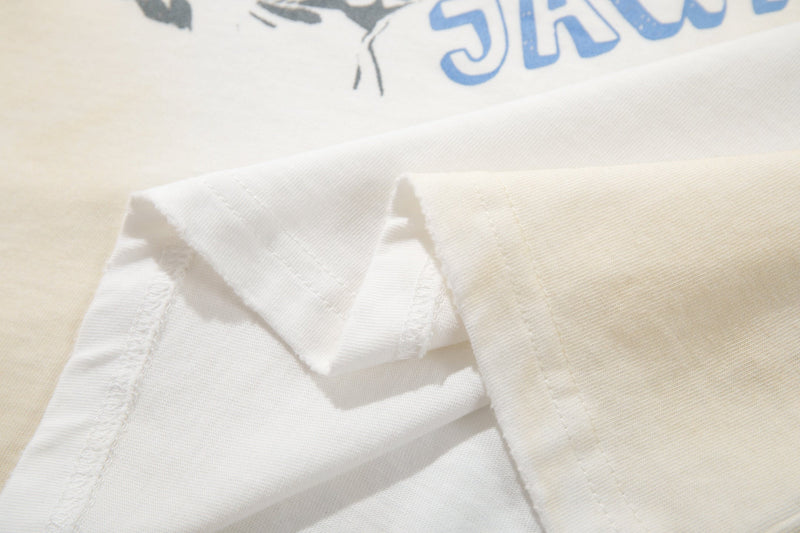 Close-up of a cream graphic tee with folded white hem and blue retro collage lettering on soft cotton fabric