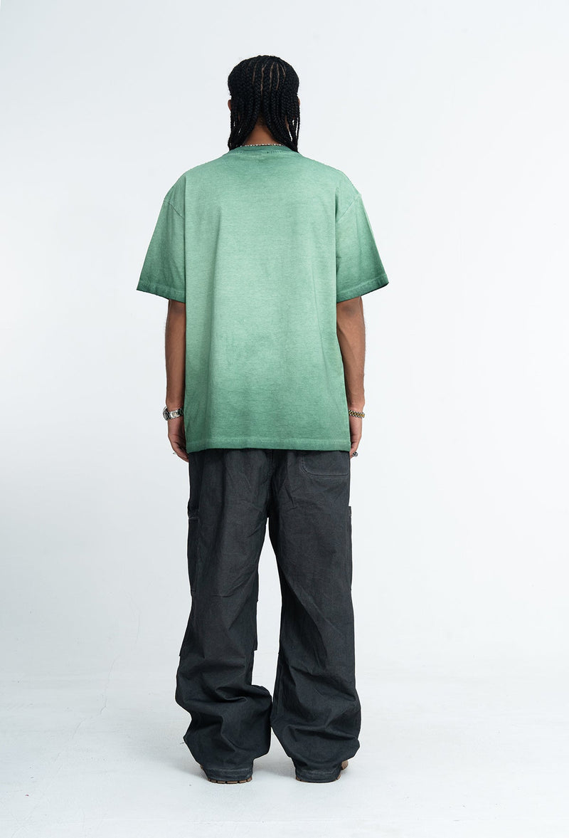 Back view of a man wearing a vintage wash green oversized graphic tee with loose black pants on a white background